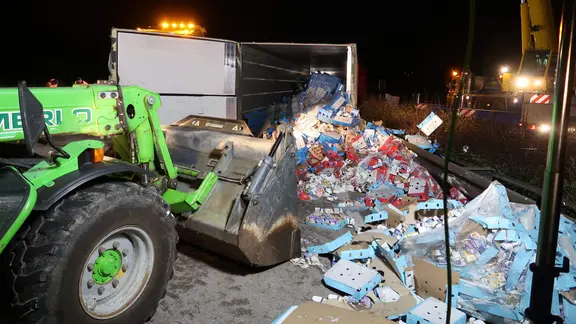 Bei nächtlichen Aufräumarbeiten nach einem Lkw-Unfall beseitigen Einsatzkräfte mit schwerem Gerät eine auf die Fahrbahn gestürzte Ladung aus Kartons und verpackten Waren.
