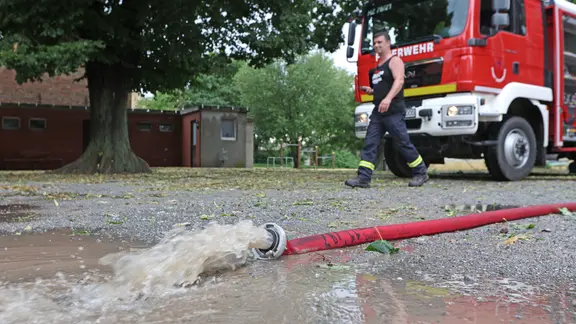 Aus einem Feuerwehrschlauch sprudelt Wasser auf die Straße, im Hintergrund das Feuerwehrauto und ein Mann