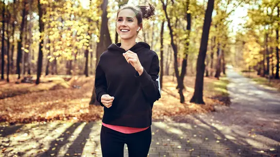 Junge Frau joggt lächelnd mit Kopfhörern durch einen Herbstwald.
