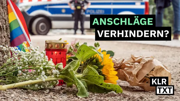Trauerkerzen, Blumen und Regenbogenflaggen liegen an einem Baum. Im Hintergrund steht Polizei.