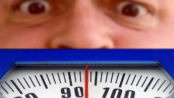 Ein Mann schaut ängstlich auf die Zahl auf der Waage. Die Waage zeigt 93 Kilogramm an. 