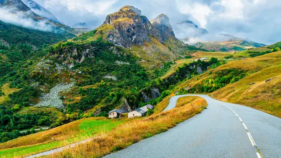 Geschwungene Landstraße führt durch eine hügelige, grün-gelbe Berglandschaft mit Felskuppen, vereinzelten Häusern und tief hängenden Wolken im Hintergrund.