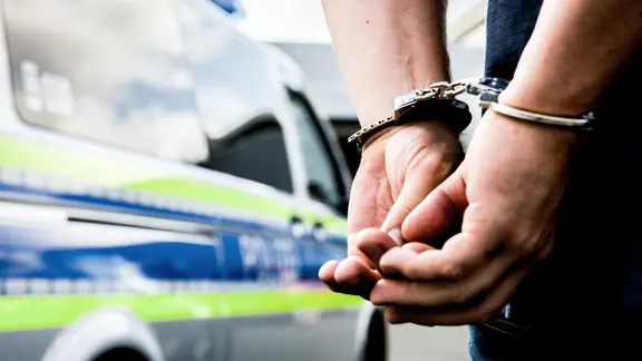 Symbolbild zu Polizeieinsatz und Strafverfolgung: Ein Mann wird mit Handschellen von der Polizei festgenommen. Im Hintergrund steht ein Streifenwagen der Polizei mit reflektierenden Markierungen.