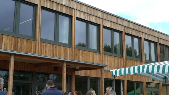 Modernes Gebäude mit Holzfassade und großen Fensterfronten. Vor dem Eingang stehen mehrere Menschen unter einem gestreiften Sonnenschutz bei einer Veranstaltung im Freien.