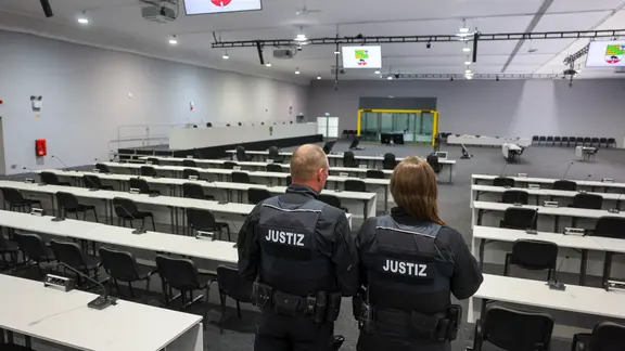 Zwei Justizbeamte stehen im Verhandlungssaal im Interims-Gerichtsgebäude für den Prozess gegen den Todesfahrer vom Magdeburger Weihnachtsmarkt, im Hintergrund eine schusssichere Glaskabine für den Angeklagten. Der Hauptsaal in der Leichtbauhalle bietet mit 1.900 Quadratmetern Platz für etwa 700 Personen.