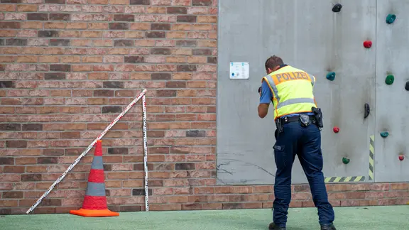 Ein Polizist misst neben einem roten Warnkegel mit einem großen Zollstock die Unfallstelle auf einem Schulhof aus