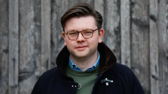 Porträt eines jungen Mannes vor einer Holzwand. Er blockt direkt in die Kamera und trägt zur Seite gekämmte  Haare, eine gerahmte Brille und einen grünen Pullover mit blauem Hemdkragen, darüber eine offene dunkle Wolljacke mit Stehkragen
