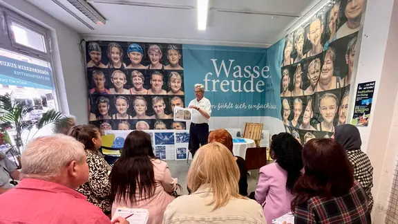 Erwachsene Menschen sitzen in einem Raum, an dessen Wand viele Gesichter abgebildet und das Wort "Wasserfreude", und tragen etwas auf Zettel ein. Vor ihnen steht ein Mann und spricht.