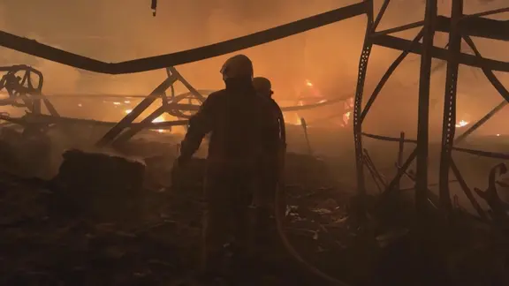 Feuerwehrmänner in Schutzhelmen zwischen Trümmern und Flammen eines Großbrandes in der Nacht.
