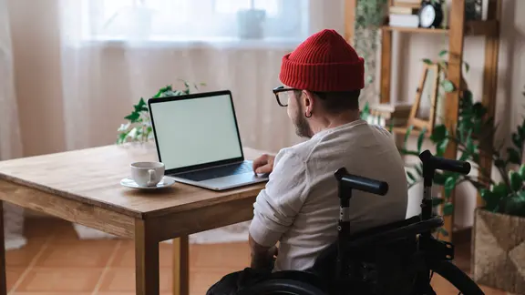 Symbolfoto: Ein Mann im Rollstuhl sitzt an einem Schreibtisch mit Laptop.