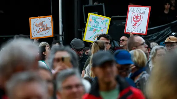 Menschen haben sich auf dem Marktplatz zur Open-Air-Veranstaltung "Halle steht auf. Gemeinsam für Demokratie. Zusammenhalt. Sachsen-Anhalt." versammelt und halten Schilder mit der Aufschrift "wir wählen", "Hass ist keine Meinung" und "Halle steht auf für Demokratie".