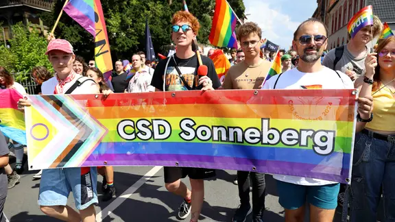 Menschen mit Regenbogenfahnen und einem Banner "CSD Sonneberg laufen über eine Straße.