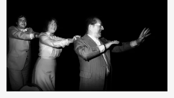 Zwei Männer im Anzug und eine Frau machen eine Polonaise, halten sich an den Schultern des Vordermanns fest