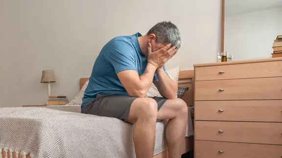 Ein Mann sitzt auf dem Bett, hält sich die Hände vor das Gesicht und zeigt Anzeichen von Verzweiflung, Verlust und psychischen Problemen.