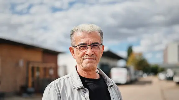 Hamdi Kayisi ist ein älterer Mann mit grauem Haar und einer hellgrauen Jacke, der in einem Gewerbegebiet steht.