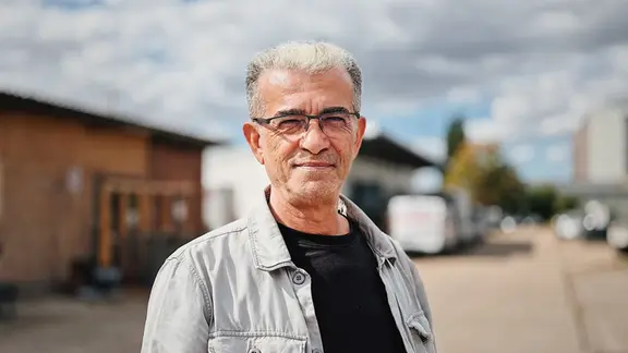 Hamdi Kayisi ist ein älterer Mann mit grauem Haar und einer hellgrauen Jacke, der in einem Gewerbegebiet steht.