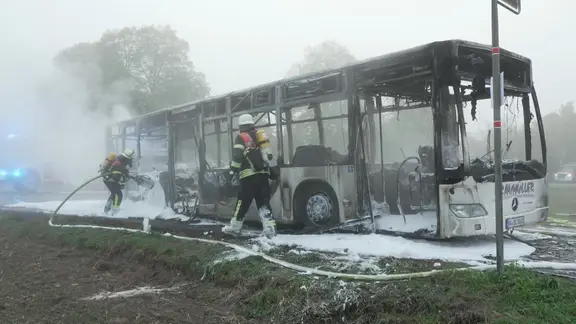 Ausgebrannter Linienbus, zwei Feuerwehrleute löschen mit Schaum.