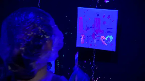 Ein Kind mit Haarschutz malt in einem Raum mit Schwarzlicht auf einer kleinen quadratischen Leinwand mit fluoreszierenden Farben.
