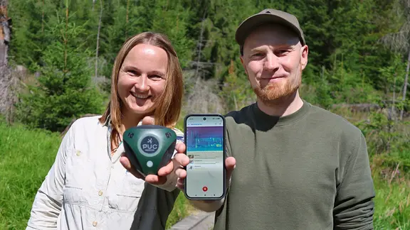 Frau mit schulterlangen blonden Haaren und Mann mit rötlichem Bart und Kappe auf Waldlichtung. Frau hält Audiologger-Gerät in die Kamera, Mann ein Smarphone mit der Birdnet-App.