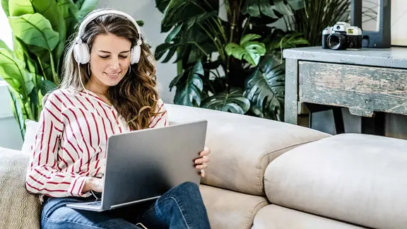 Eine lächelnde Frau mit Laptop und Kopfhörern sitzt auf einer Couch