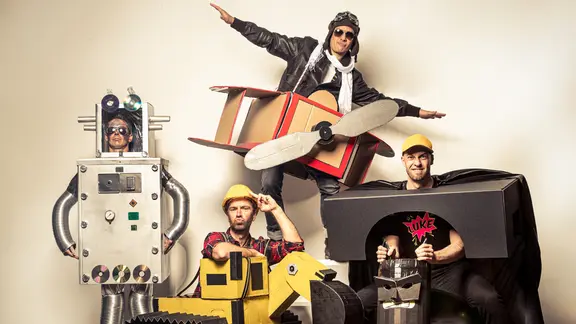 Maybebop: Inszeniertes Bandfoto zeigt vier Männer in Pappkostümen, ein Roboter, ein Bagger, ein Propellerflugzeug und ein Batman
