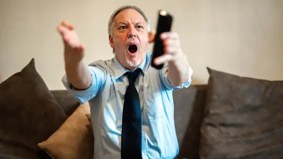 Ein Mann sitzt auf einem Sofa, hat eine Fernbedienung in der Hand und schreit wütend.