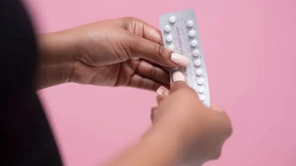 Nahaufnahme zwei Hände vor einem einheitlich rosa Hintergrund. Eine Person hält mit beiden Händen eine silberfarbene Blisterpackung mit weißen, filmbeschichteten Tabletten (Antibabypille), wobei ein Finger eine Tablette aus der Verpackung drückt. Auf der Person und im Bild ist ansatzweise ein dunkler Kleidungsausschnitt am linken Bildrand zu erkennen.