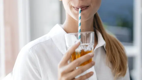 Junge Frau mit blondem Pferdeschwanz in weißer Bluse trinkt Tee aus einem Glas mit blau-weiß gestreiftem, mutmaßlichem Papierstrohhalm in einem hellen Raum, blaue Wand mit weißem Regal unscharf im Hintergrund.