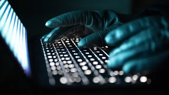 Eine Person tippt mit behandschuhten Händen auf einer beleuchteten Laptop-Tastatur. 