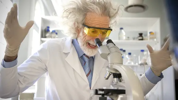Ein älterer Professor mit zerzaustem, weißem Haar und Bart trägt einen weißen Laborkittel, eine blaue Krawatte und eine gelb getönte Schutzbrille. Er steht in einem hellen Labor und beugt sich überrascht, mit weit aufgerissenem Mund und erhobenen Händen, über ein Mikroskop, während im Hintergrund Laborausrüstung auf Regalen zu sehen sind.