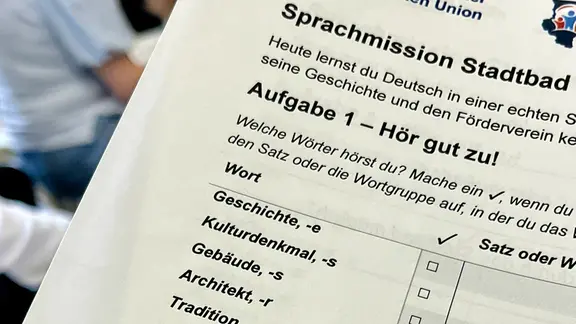 Ein Zettel mit einer Tabelle, über der steht: "Sprachmission Stadtbad" und "Aufgabe 1: Hör gut zu!"