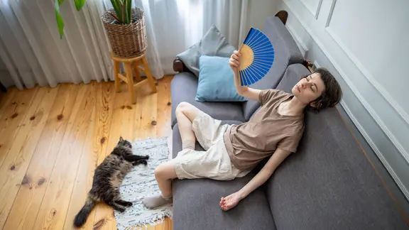 Eine Person liegt mit einem Fächer auf einem Sofa, daneben liegt eine Katze