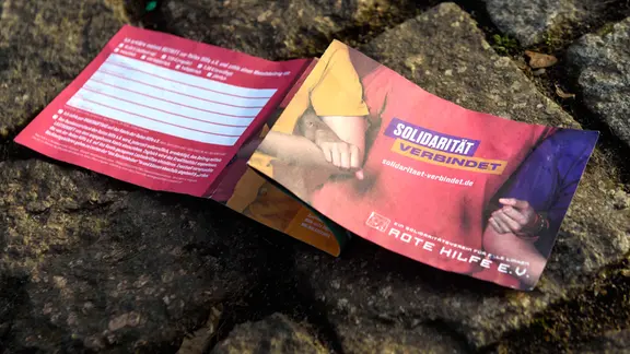 Auf Kopfsteinpflaster liegt ein aufgeklappter Flyer des Vereins "Rote Hilfe" mit der Aufschrift "Solidarität verbindet"