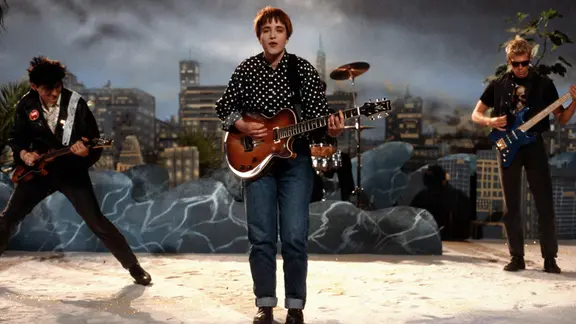 Eine Frau mit Gitarre steht auf einer Bühne; hinter ihr sind zwei weitere Musiker mit Gitarren und die Silhouette einer Großstadt zu sehen.