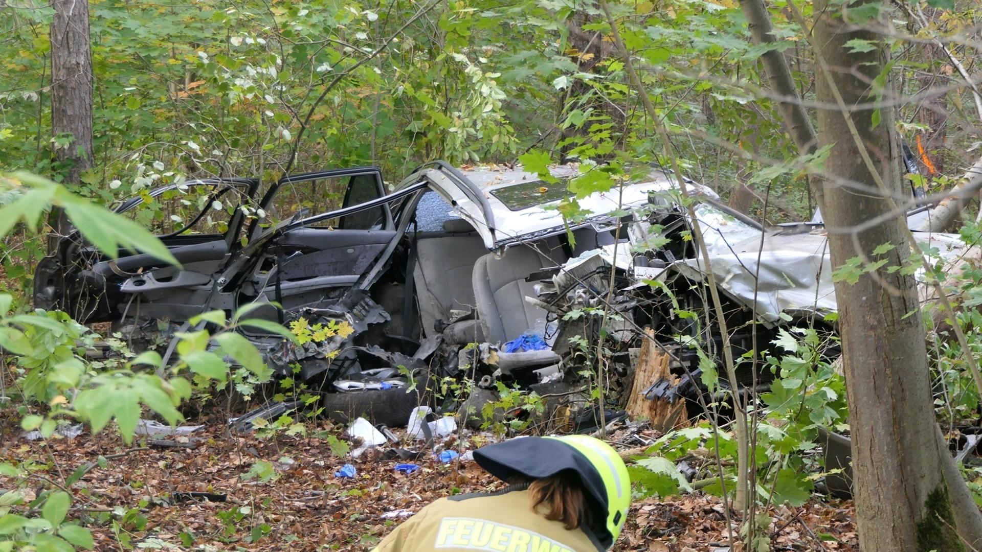 Ein völlig zerstörtes Auto liegt in einem Waldstück. Ein Feuerwehrmann ist von hinten zu sehen. | Medienportal Grimma/Sören Müller