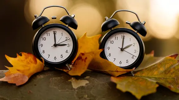 Zwei Wecker auf Herbstlaub, einer zeigt drei Uhr, der zwei Uhr.