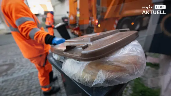 Eine mit Plastiküll und Fremdstoffen falsch befüllte Biomülltonne steht in der Innenstadt, dahinter leeren Mitarbeiter die Biomülltonnen. 