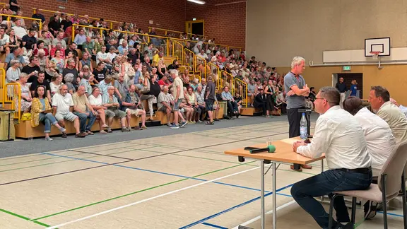 Menschen sitzen in einer Turnhalle und schauen auf einen Tisch mit Menschen