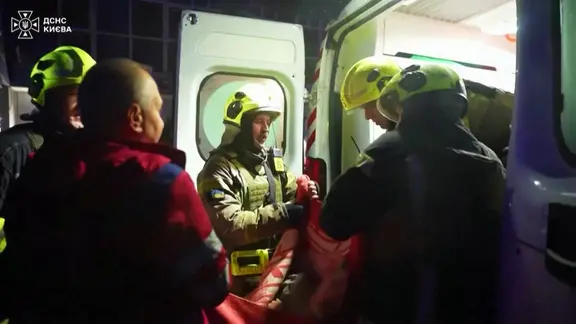 Mehrere Reetungshelfer mit Uniformen und Helmen heben eine verletzte Person in einen Krankenwagen.