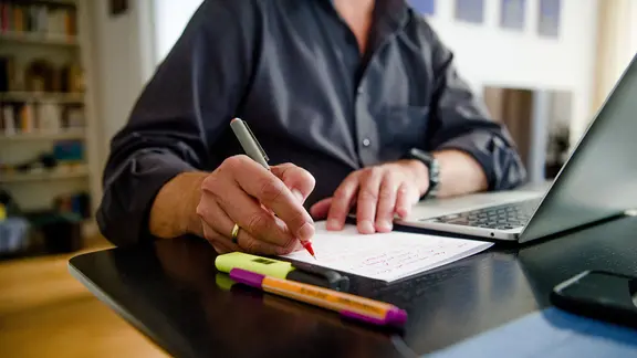 Ein Mann sitzt zuhause an einem Esstisch. Er schreibt mit einem Stift auf ein Blatt Papier neben einem Laptop.