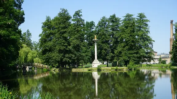 Ein Stadtteich mit Insel, Holztreppe und Säule liegt idyllisch in einem Park. 