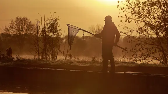 Ein Fischer steht mit leerem Netz im Sonnenuntergang an einem Teich