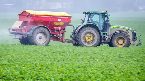 Ein Traktor fährt mit einem Streuwagen über ein Feld und bringt Dünger aus. 