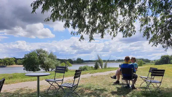 An einem Fluss auf einer Wiese unter einem Baum sitzen zwei Menschen auf Stühlen an einem kleinen Tisch
