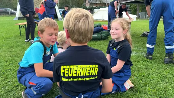 Mehrere Kinder in Einsatzkleidung der Freiwilligen Feuerwehr sitzen auf einer Wiese zusammen