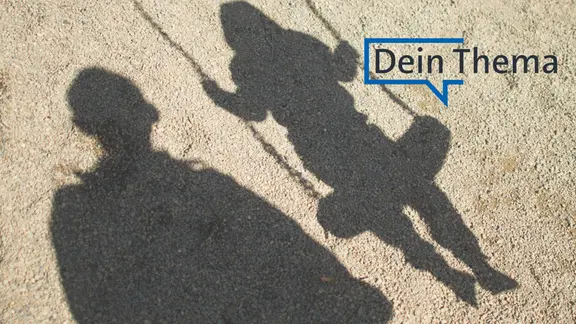 Der Schatten von einem Mann und einem schaukelnden Flüchtlingsjungen fallen auf Sand auf einem Spielplatz