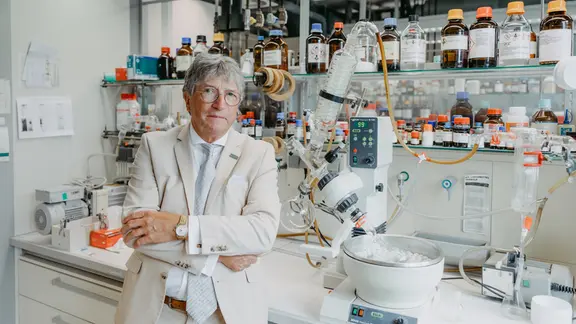 Ein Mann mit grauen Haaren und Brille steht in einem Chemielabor vor einer Vielzahl von Glasflaschen, Geräten und Schläuchen. Er trägt einen hellen Anzug und lehnt mit verschränkten Armen an einem Labortisch.