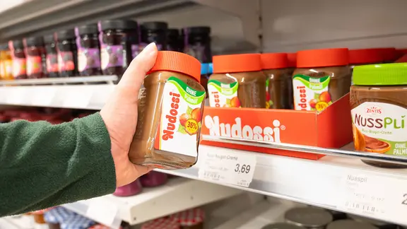 Eine Hand hält vor einem Supermarktregal ein Nudossi-Glas