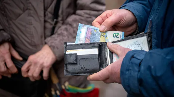 Eine ältere Person hält im öffentlichen Raum eine geöffnete Geldbörse in der Hand, aus der ein 20-Euro-Schein hervorschaut.