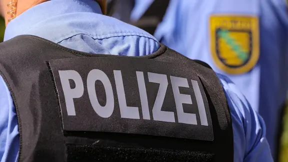 Aufschrift Polizei auf Polizeiweste im Anschnitt. Im Bildhintergrund das Landeswappen Polizei Sachsen in der Bildunschärfe.